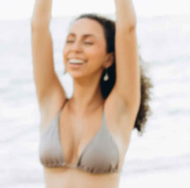 Mulher de biquíni na praia, braços pra cima, rindo, com o mar claro ao fundo