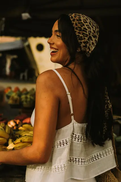 Mulher de lenço de oncinha no cabelo rindo, de costas, numa feira de frutas
