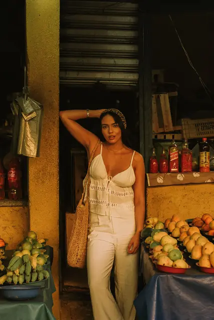 Mulher de conjunto branco encostada numa porta, entre bancas de frutas
