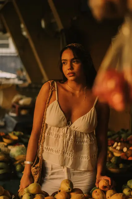 Mulher de blusa branca rendada numa feira, entre caixas de frutas, luz baixa