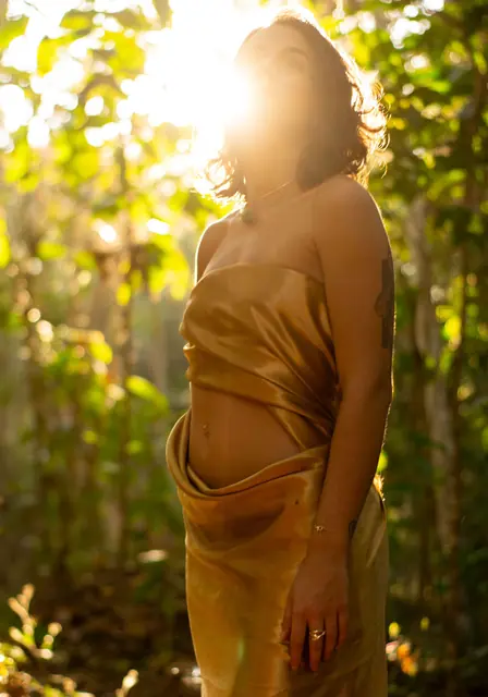 Mulher de vestido acetinado dourado entre plantas, com o sol por trás
