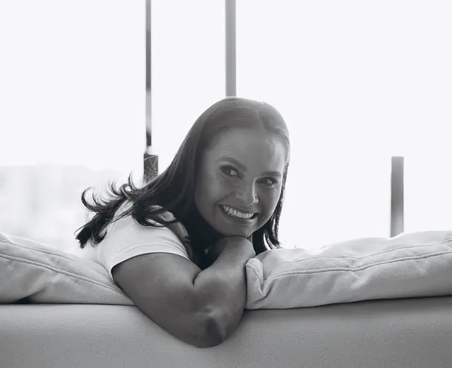 Mulher sorrindo, deitada de bruços num sofá branco, em preto e branco