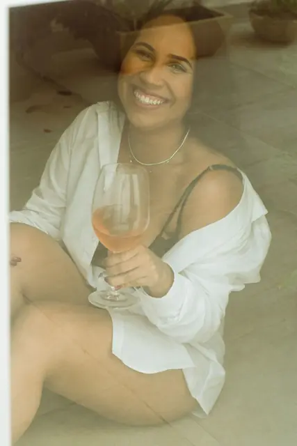 Mulher de camisa branca aberta sentada no chão, sorrindo, com uma taça de vinho na mão