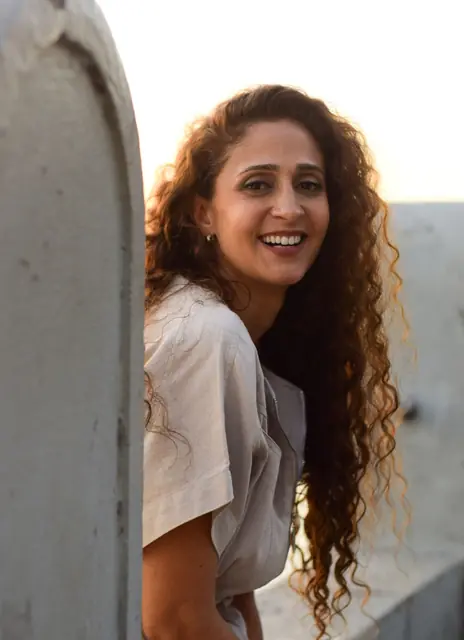 Mulher de cabelo longo e cacheado sorrindo, encostada numa parede clara, luz de fim de tarde