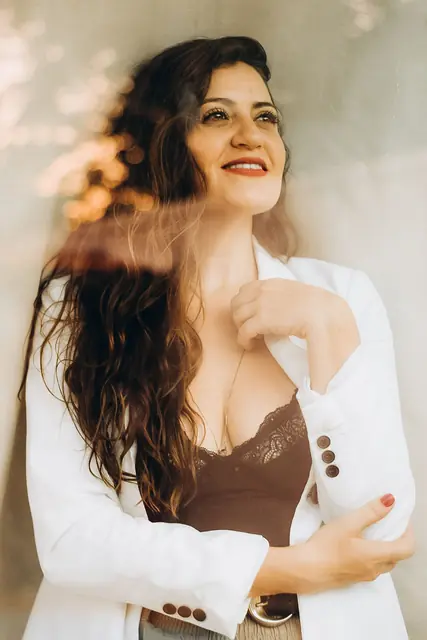 Mulher de cabelo longo e cacheado, de blazer branco, sorrindo com a mão no peito