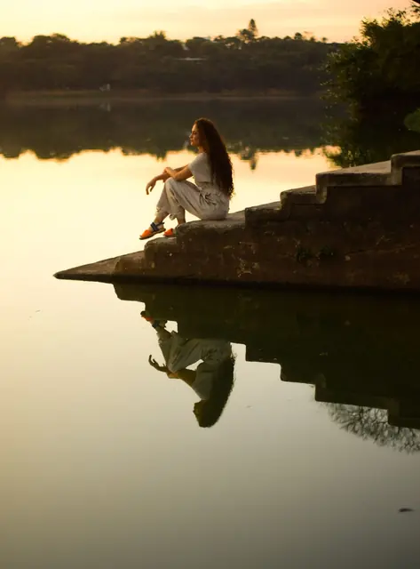 Mulher sentada na beira de um lago no fim da tarde, com o reflexo na água